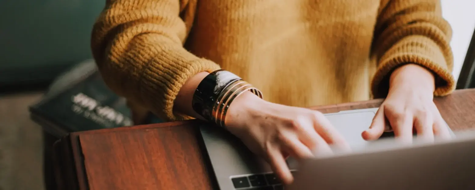 Eine Person mit einem senfartigen Pullover tippt auf einer Laptop-Tastatur. Ihr Handgelenk ist mit mehreren Armreifen geschmückt. Der Hintergrund zeigt unscharfe Gegenstände, die nicht im Fokus stehen.