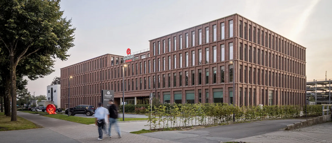 A modern brick building with multiple windows and an illuminated logo on the roof. In front, two people walk along a pathway, and a red sculpture is visible on the left. Surrounding greenery adds to the urban landscape.