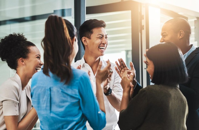 A diverse group of six people stands in a bright office space, laughing and clapping while engaging in a joyful conversation. Their expressions convey excitement and camaraderie, showcasing a positive and friendly atmosphere.