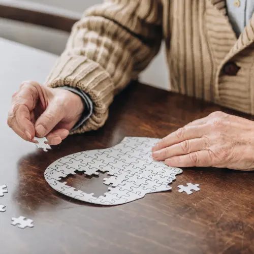 An older person’s hands are seen putting together a white jigsaw puzzle on a wooden table. A few puzzle pieces lie scattered nearby. The scene captures the focus and concentration involved in completing the puzzle.