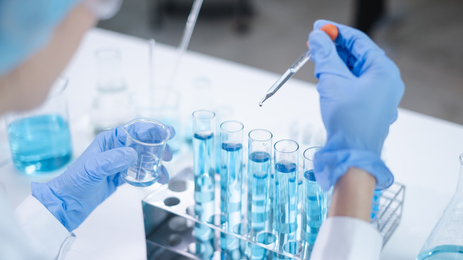A scientist wearing gloves and a lab coat is holding a small glass beaker in one hand and a dropper in the other, preparing to add a liquid to a rack of test tubes filled with a blue solution.