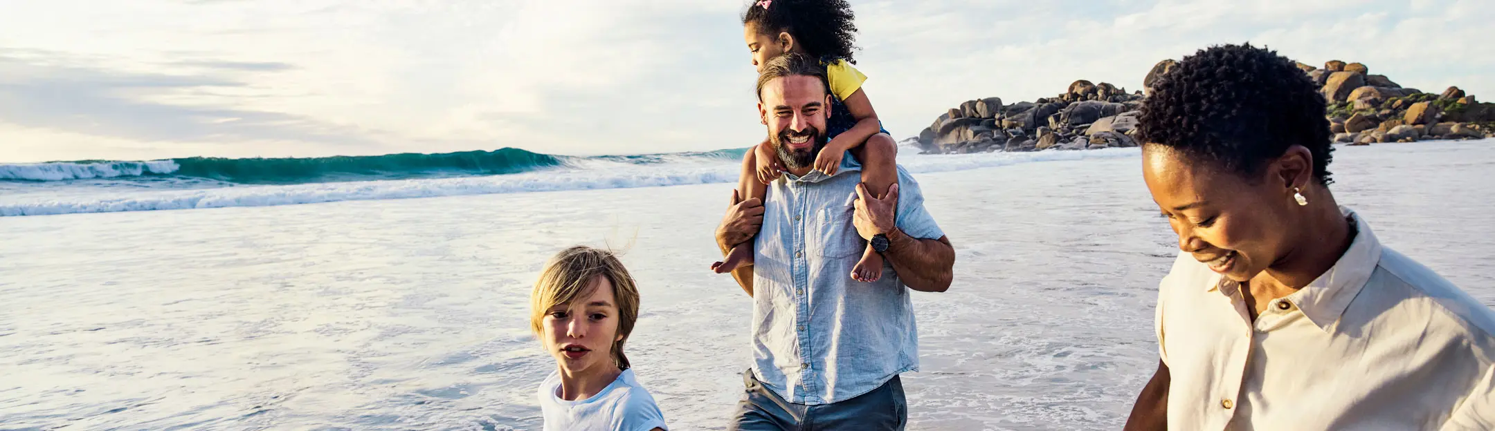 A family walks along a beach during sunset. A man carries a girl on his shoulders, while two other children walk beside them. The ocean waves crash in the background, with rocky formations visible in the distance.