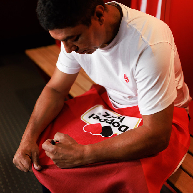 A man is seated on a bench in a locker room, wearing a white t-shirt and focused on adjusting a red towel with a graphic design. The setting has wooden benches and red walls, suggesting a sports environment.