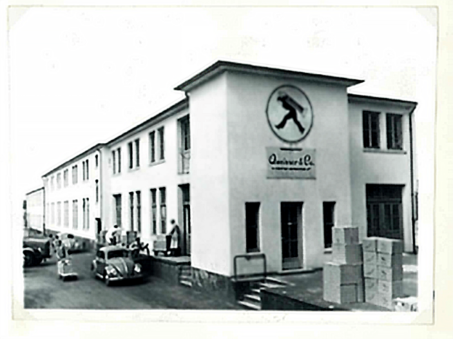 Black and white photo of a large building with a rounded logo featuring a person walking. The front entrance has stairs, and several people are seen near parked cars and stacked boxes in front of the building. 