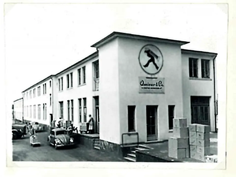 Black and white photo of a large building with a rounded logo featuring a person walking. The front entrance has stairs, and several people are seen near parked cars and stacked boxes in front of the building. 