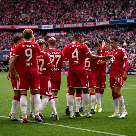 A group of FC Bayern Munich players in red uniforms huddle together, celebrating and interacting on the field. The background features a cheering crowd, indicating a football match atmosphere. The players show camaraderie and enthusiasm.
