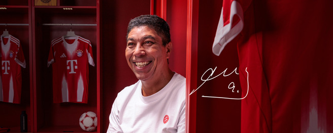 A smiling man sits inside a red locker room, featuring FC Bayern Munich jerseys in the background. A soccer ball is nearby, and there are team colors and memorabilia visible. The image conveys a sense of warmth and pride associated with the team.
