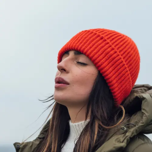 A woman with long, dark hair is wearing an orange beanie and a cozy, green jacket. She has her eyes closed and appears to be enjoying a moment of tranquility outdoors. The background is softly blurred, suggesting an open sky.