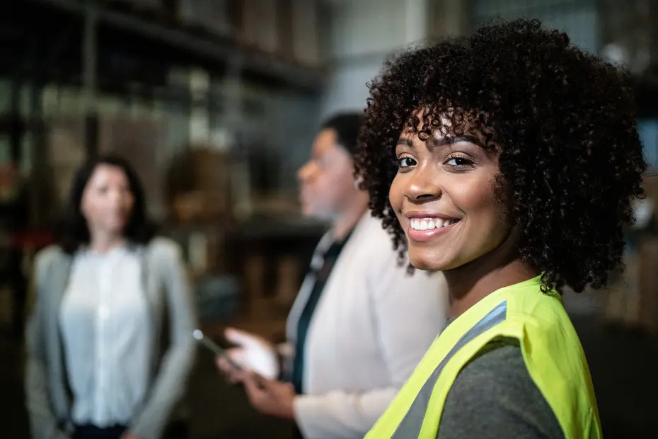 A smiling woman with curly hair wearing a yellow safety vest is in focus, while two other women are blurred in the background. They appear to be in a warehouse setting, discussing or interacting with each other. The atmosphere suggests a work-related environment.