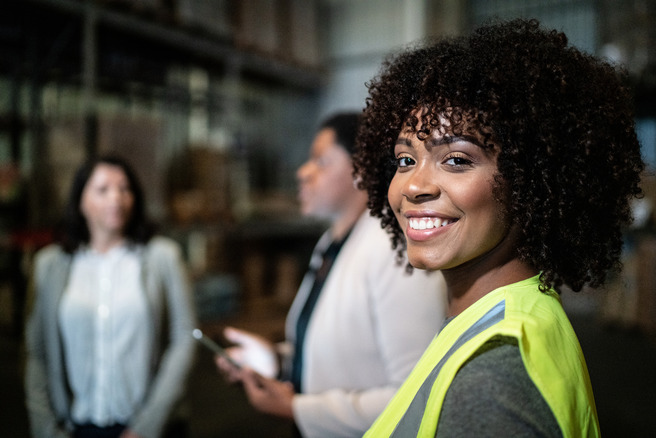 A smiling woman with curly hair wearing a yellow safety vest is in focus, while two other women are blurred in the background. They appear to be in a warehouse setting, discussing or interacting with each other. The atmosphere suggests a work-related environment.