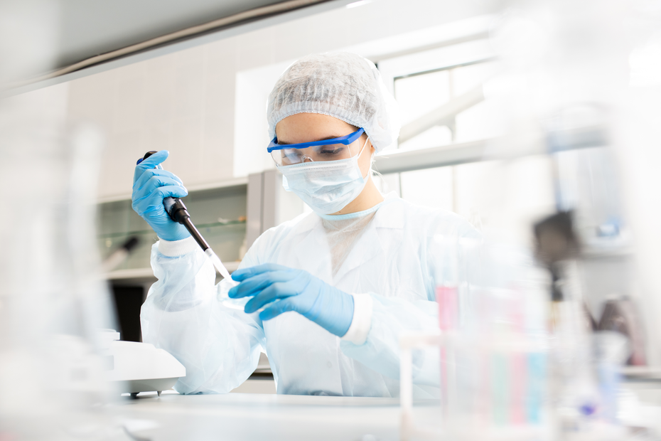 A laboratory technician in a white coat, cap, and protective gear is using a pipette to measure a liquid. The background features laboratory equipment, emphasizing a scientific research setting.
