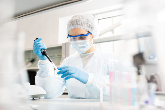 A laboratory technician in a white coat, cap, and protective gear is using a pipette to measure a liquid. The background features laboratory equipment, emphasizing a scientific research setting.
