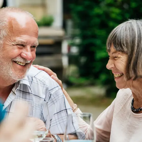 A happy elderly couple share a joyful moment together, smiling and laughing at each other. They are seated at a table, with glasses nearby, enjoying their time in an outdoor setting.