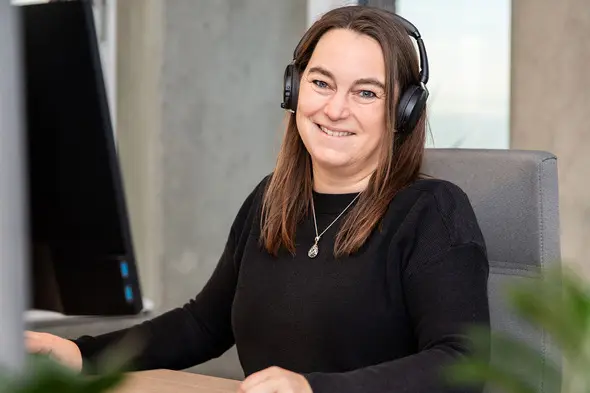 A woman wearing headphones smiles while sitting at a desk in front of a computer monitor. She has long brown hair and is dressed in a black sweater. The setting appears to be a modern office with a neutral background.