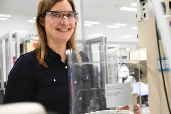 A smiling female scientist wearing safety glasses stands in a laboratory environment, operating equipment. She appears engaged and friendly, with a clean and organized workspace in the background.