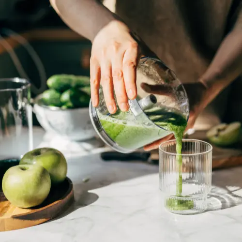 A person is pouring green smoothie from a blender into a clear glass. Fresh green apples and cucumbers are visible on a wooden cutting board and in the background. The setting is bright, suggesting a healthy and refreshing drink preparation.