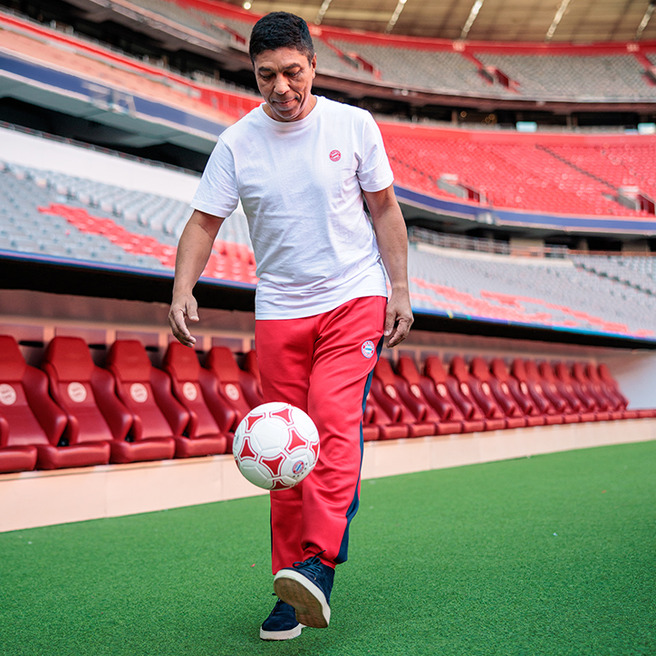A person in a white shirt and red pants is skillfully juggling a soccer ball on a green turf field inside a stadium. Red seats are visible in the background, suggesting it is a sports venue. The atmosphere conveys a sense of playfulness and athleticism.