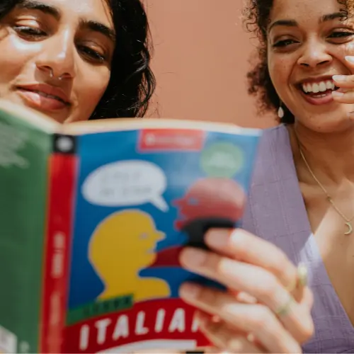 Two women are joyfully engaged in reading a colorful language book titled "Learn Italian." One woman holds the book up, while the other smiles, looking at it. Their expressions convey excitement and enjoyment in learning together. The background features a warm, neutral tone.