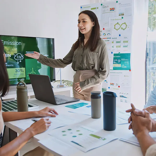 A woman stands presenting in front of a screen displaying emission data, while seated individuals observe and engage in a meeting. The table has laptops and water bottles, and the backdrop features various informational posters related to sustainability.