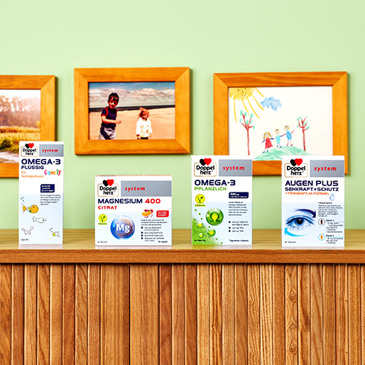 Four product boxes are displayed on a wooden shelf against a light green wall. The products are labeled Omega-3, Magnesium 400 Citrate, and Augen Plus. In the background are framed photographs and children's drawings, adding a vibrant atmosphere.