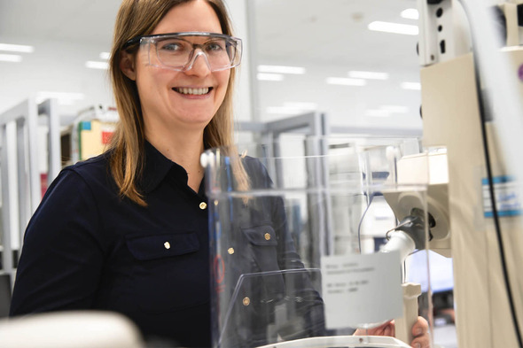 A smiling female scientist wearing safety glasses stands in a laboratory environment, operating equipment. She appears engaged and friendly, with a clean and organized workspace in the background.