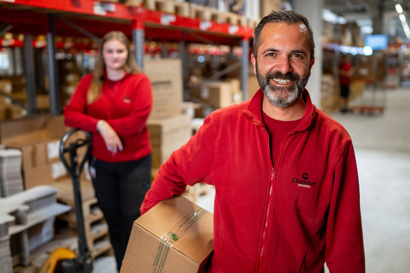 A smiling man in a red uniform holds a cardboard box in a warehouse. Behind him, a woman in a matching red uniform stands near a pallet jack, both present in a setting filled with stacked boxes. The environment appears busy and organized.
