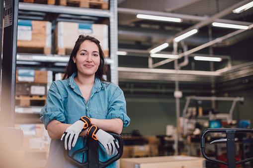 A woman stands confidently with her hands on a pallet jack in a warehouse setting. She is wearing a denim shirt and work gloves, with shelves of boxes in the background, creating a busy industrial atmosphere.