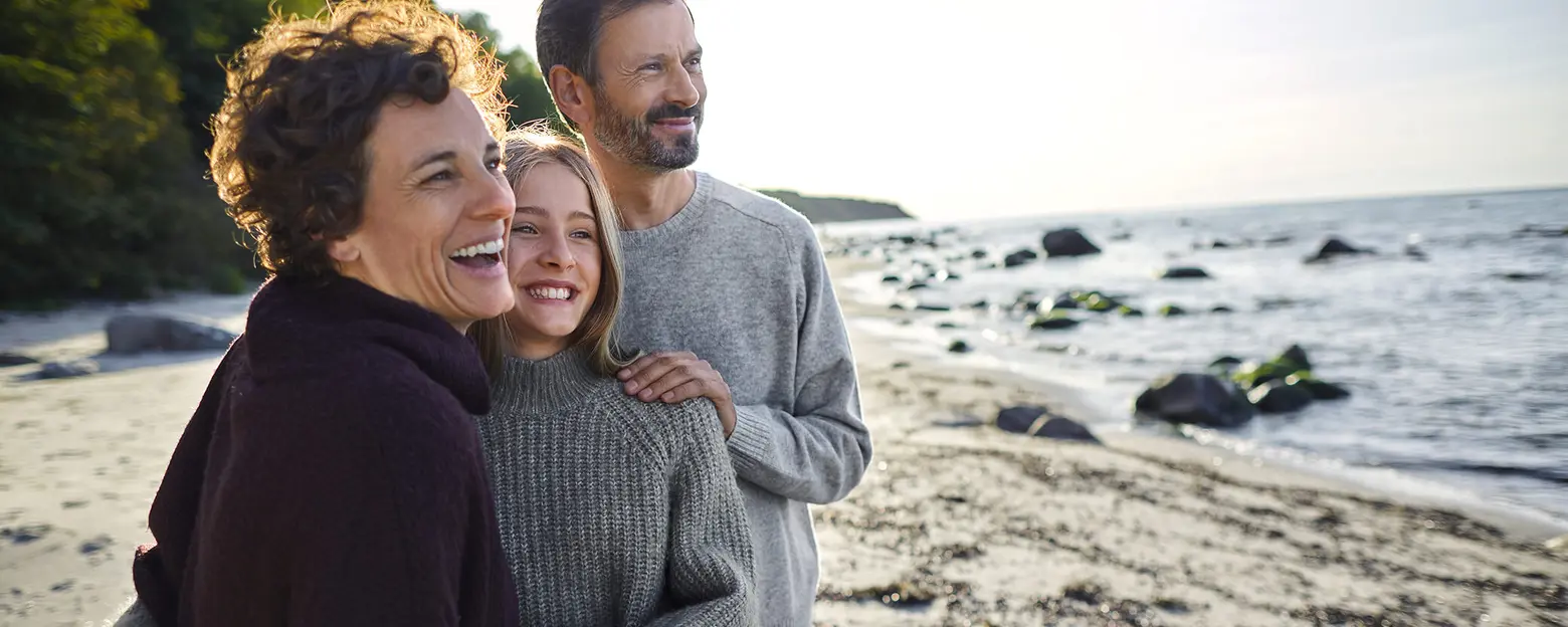 Three people stand together on a beach, smiling and embracing each other. They appear happy and relaxed, with the ocean and a rocky shoreline in the background. The scene conveys warmth and connection among the individuals.